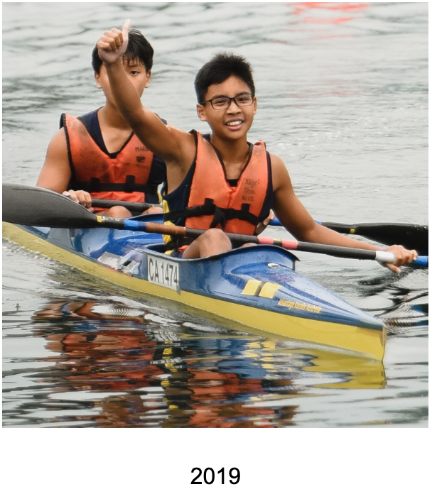 Two boys in orange life vests paddle a blue and yellow kayak, one giving a thumbs up, in 2019.